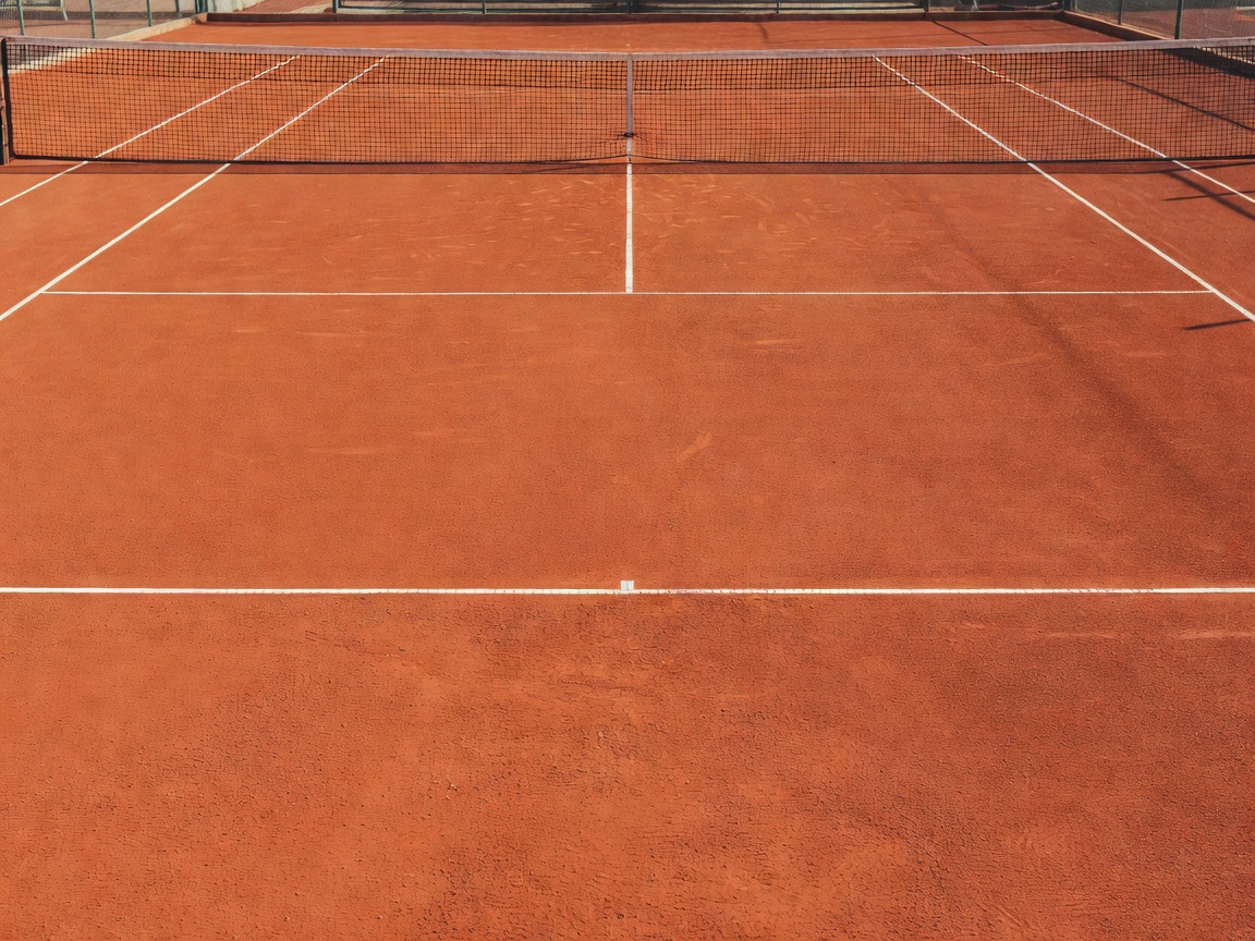 A public red clay tennis court bathed in natural sunlight with crisp white lines